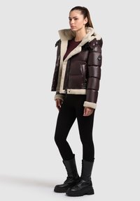 Giacca imbottita marrone con colletto in finta pelliccia, chiusura con zip e tasche laterali, abbinata a leggings neri e stivali neri fino al ginocchio.
