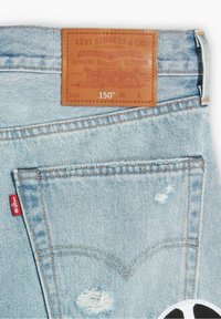 Jeans de mezclilla de color azul claro con un parche de cuero con texto en relieve, costuras visibles y detalles desgastados cerca de los bolsillos.