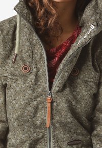 Grön jacka med blommönster, tillverkad av texturerat tyg, med dragkedjestängning, två bröstknappar och en läderdragre.