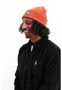 Orange ribbed beanie with a black logo patch, worn with a black corduroy button-up shirt featuring a chest pocket and subtle branding details.