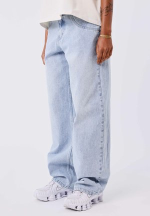 Helle blaue, weite Denim-Jeans mit strukturierter Oberfläche und sichtbaren Nähdetails. Kombiniert mit weißen Sneakern mit auffälligem Design.