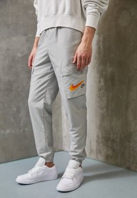 Šedé cargo kalhoty s oranžovým logem Nike, mají boční kapsy a elastické manžety. Nosené s bílými teniskami na texturovaném pozadí.