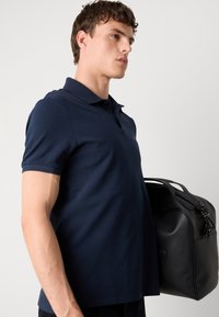 Ung mand i navyblå polo shirt bærer en sort læder rejsetaske, set fra siden mod en ensfarvet lys baggrund.