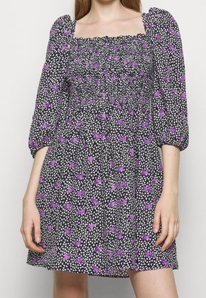 Czarna sukienka z fioletowym kwiatowym wzorem, marszczony kwadratowy dekolt, bufiaste rękawy do połowy przedramienia, noszona przez osobę z rękami swobodnie opuszczonymi wzdłuż boków.