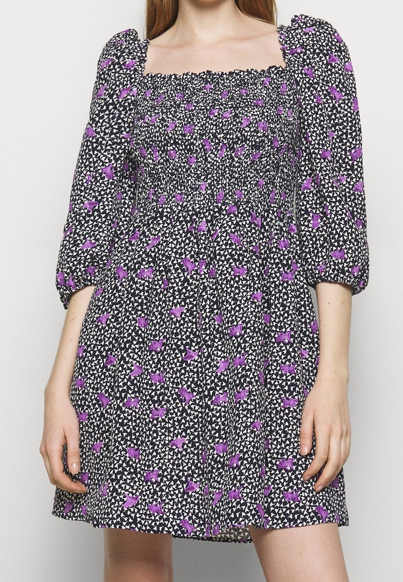 Robe noire avec motif floral violet, encolure carrée froncée, manches trois-quarts bouffantes, portée par une personne les mains détendues le long du corps.
