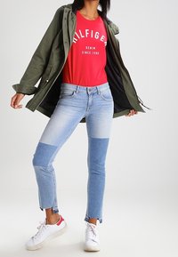 Person som bär tvåfärgad ljus- och mörkblå jeans, en röd t-shirt med grafik, en grön jacka och vita sneakers med röda accenter.