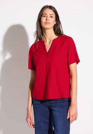 Rode blouse met korte mouwen en een V-hals, gemaakt van een soepele stof en met een losse pasvorm. De zoom valt onder de taille en is gecombineerd met donkerblauwe jeans.