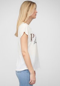 T-shirt blanc à manches courtes avec un motif graphique en texte léopard. Tissu doux, coupe décontractée, vue de côté sur un fond uni.