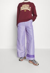 Kastanjebrun sweatshirt med "ADIDAS ORIGINALS" logotyp i gult. Vida ljuslila byxor med lila tre-randsaccent vid fållen. Skor i blandade material.