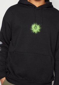 Sudadera negra con un bolsillo frontal, que presenta un vibrante diseño gráfico en verde de un corazón con acentos de estallido. Cuerda ajustable en la capucha.