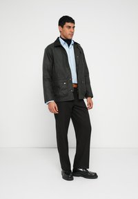 Donkergroene jas met ritssluiting aan de voorkant, twee lage zakken en een gestructureerde afwerking. Draagt over een lichtblauw overhemd en zwarte broek, gecombineerd met zwarte schoenen.