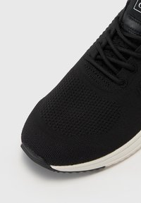 Pantof black pentru sport cu un superior texturat, având un design din plasă. Talpa este albă și are o formă ușor curbată. Sireturi sunt negre.