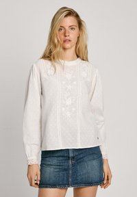 Blusa bianca ricamata con maniche lunghe, pois a trama, e dettagli in pizzo al colletto e ai polsini, abbinata a una mini gonna in denim.