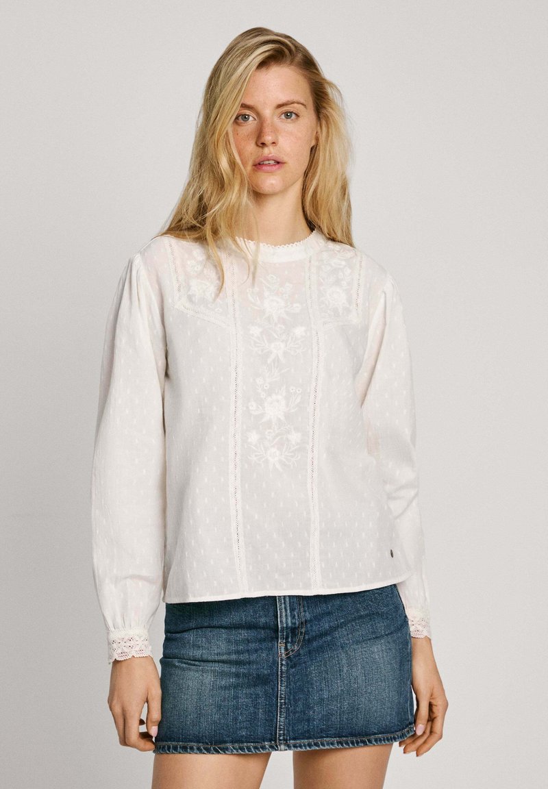 Blusa bianca ricamata con maniche lunghe, pois a trama, e dettagli in pizzo al colletto e ai polsini, abbinata a una mini gonna in denim.
