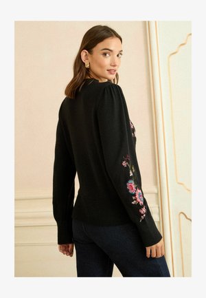 Pulover negru cu mâneci bombate și broderie florală în roz și albastru pe brațe. O margine cu dungi și o siluetă ajustată.