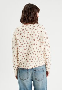 Pull crème avec des taches marron, texture tricotée, col rond et ourlet bord-côte. Associé à un jean en denim bleu clair avec des poches arrière.