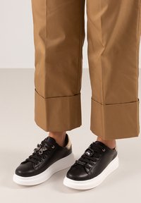 Zapatillas negras con suela blanca, que presentan detalles decorativos en los cordones y un tacón dorado texturizado. Combinadas con pantalones cortos beige.