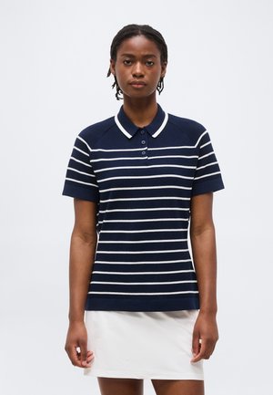 Donna con capelli corti intrecciati che indossa una polo a maniche corte a righe orizzontali blu navy e bianche e una gonna bianca.