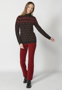 Pull en tricot noir avec motif géométrique rouge, col montant, manches longues. Assorti à un pantalon en velours côtelé bordeaux et des bottes noires à lacets.