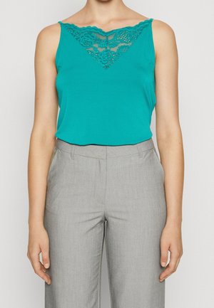 Frau trägt ärmelloses türkisfarbenes Oberteil mit Spitzenbesatz am Ausschnitt, kombiniert mit hoch taillierter hellgrauer Hose, steht vor einfachem Hintergrund.