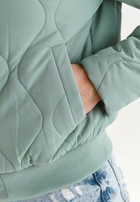 Hand in der Tasche einer hellgrünen, gesteppte Jacke mit geripptem Saum, kombiniert mit blauen, gemusterten Jeans.