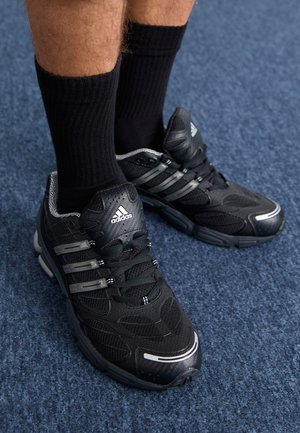 Chaussures de sport noires avec une tige en tissu texturé, des accents gris et des lacets. Portées avec des chaussettes noires côtelées sur une surface de tapis bleu.