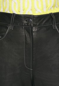 Närbild av svarta läderbyxor med vit sömnad och metallknapp, kombinerade med en gul kabelstickad tröja.