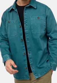 Camicia di jeans verde teal con chiusura a bottoni, due tasche sul petto e bottoni marroni. Presenta maniche lunghe e un classico design del colletto.