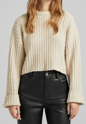 Personne portant un pull en maille côtelée beige à manches longues et un pantalon taille haute en faux cuir noir, debout devant un fond neutre.