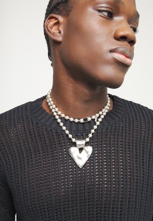 Persona de piel oscura con cabello trenzado que lleva un top de punto negro y dos collares de cuentas plateadas, uno con un colgante grande en forma de corazón brillante.