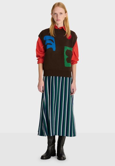 Femme portant un pull sans manches marron avec des formes colorées, une chemise rouge, une longue jupe rayée et des bottes noires, debout devant un fond uni.
