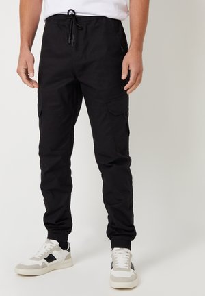 Pantaloni cargo neri con polsini elasticizzati e tasche, realizzati in un materiale leggero. Abbinati a sneakers bianche e grigie.