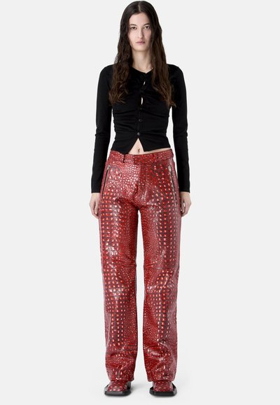 Femme portant un haut noir moulant à manches longues et un pantalon taille haute brillant à motif crocodile rouge avec des poches zippées.