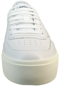 White leather sneakers with flat laces, perforated toe, thick rubber sole, and textured accents. Brand label visible on tongue.