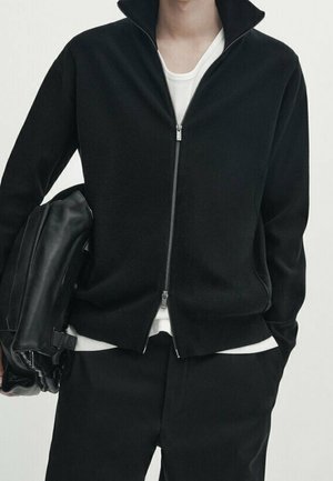 Personne portant une veste noire zippée sur une chemise blanche, un pantalon noir, tenant un sac en cuir noir, debout avec une main dans la poche.