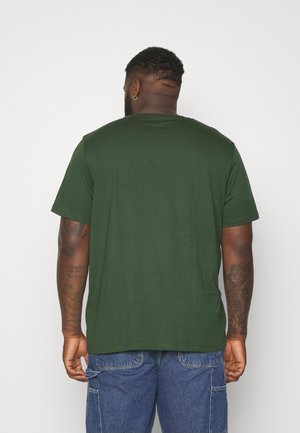 Zwarte man met tatoeages, die een donkergroen T-shirt met korte mouwen en blauwe denim cargoshorts draagt, staat met zijn rug naar een effen achtergrond.