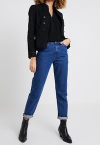 Kvinna i svart knäppt jacka, svart topp, blå upprullade jeans och svarta klackade ankelstövlar, står med ena handen i fickan.