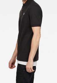 Mustat lyhythihaiset polo-paidat, joissa on kaulus, pieni logo vasemmassa rintapielessä, ja hieman kentälle kohoava malli, jossa on sileä pinta.