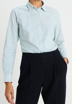 Vrouw draagt een lichtblauw-wit gestreept overhemd, gestopt in een zwarte broek met hoge taille, met één hand in de zak.