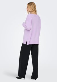 Liliowy, oversize sweter z prążkowaną fakturą i bocznymi rozcięciami, w połączeniu z czarnymi spodniami o szerokich nogawkach oraz czarnymi butami na obcasie.