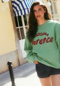 Lys grøn sweatshirt med teksten "Missing Florence" i rød, parret med mørke denim shorts. Blødt stof, afslappet pasform, synlig syning.