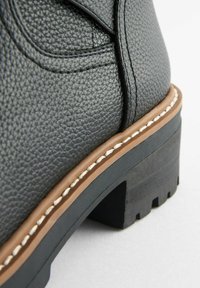 Schwarze Lederstiefel mit einer strukturierten Oberfläche, kontrastierender brauner Sohle und weißen Nähten. Robuster Absatz mit einem groben Profil.