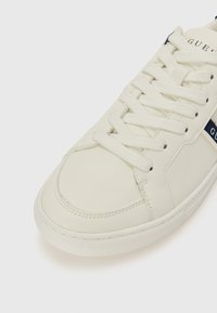 White leather sneaker with textured surface, flat laces, and a blue accent stripe featuring the brand name. Rounded toe and rubber sole.