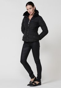 Chaqueta de plumas negra con alto cuello, cierre de cremallera y paneles laterales texturizados. Combinada con pantalones negros de cuadros ajustados y zapatos planos.