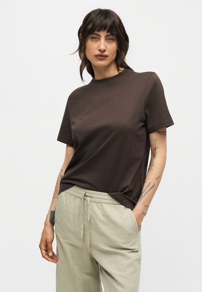 Γυναίκα με σκούρα μαλλιά και τατουάζ, που φοράει καφέ απλό κοντομάνικο μπλουζάκι και ανοιχτό μπεζ παντελόνι με κορδόνι, με τα χέρια στις τσέπες.