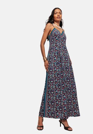 Femme portant une robe longue sans manches à motifs avec de fines bretelles, accessoirisée d'un bracelet, d'un collier, de boucles d'oreilles et de sandales noires à talons hauts.