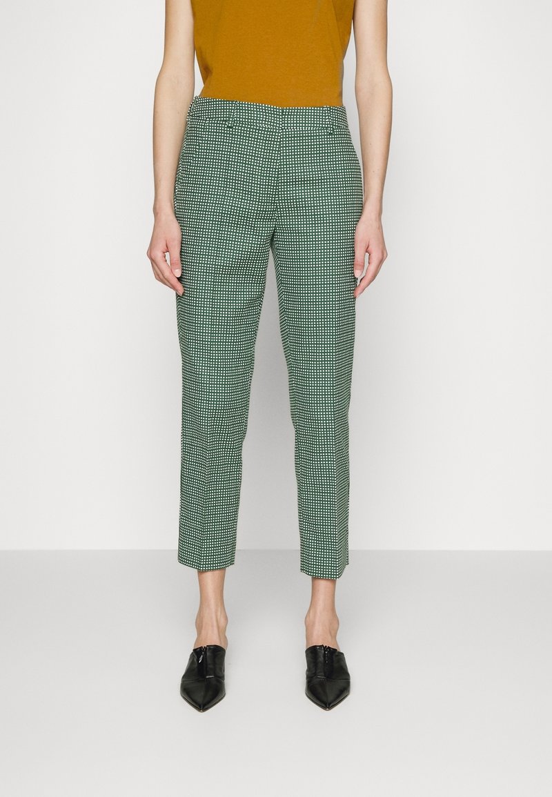 Femme portant un pantalon court à petits carreaux verts et blancs, un haut sans manches moutarde et des chaussures pointues noires, debout sur fond blanc.