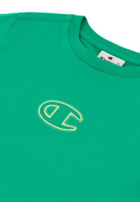 Camiseta de algodón verde con cuello redondo, que presenta un logo bordado amarillo en el pecho. Textura suave con mangas cortas.