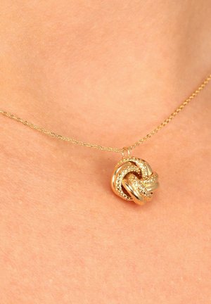 Goldene Halskette mit einem strukturierten Knoten-Anhänger und ineinander verwobenen Strängen, vor einem hautfarbenen Hintergrund. Die Kette hat ein ovales Glieddesign.