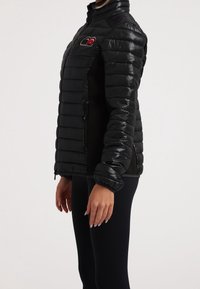 Personne portant une veste matelassée noire cintrée avec des poches zippées sur les côtés et un petit patch rouge et blanc sur la poitrine, associée à des leggings noirs.
